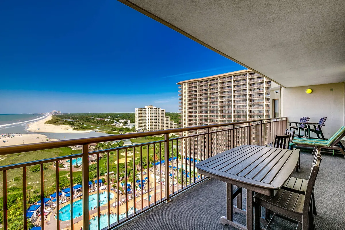 An exterior view of a private balcony furnished with comfortable seating, offering a sweeping ocean view. Below, the resort pool and sandy beach are visible, inviting relaxation and social gatherings. The tranquil water and open sky evoke a sense of leisure and exclusivity.