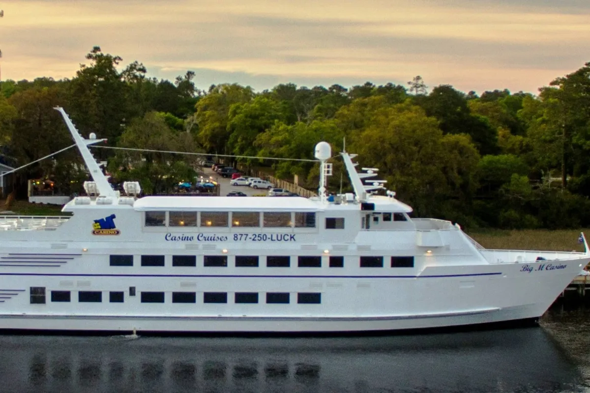This exterior shot highlights a large white cruise ship docked amid coastal greenery, with casino branding visible. The festive mood evokes the sense of adventure and special experiences available to guests, such as sunset cruises on the Barefoot Queen Riverboat.