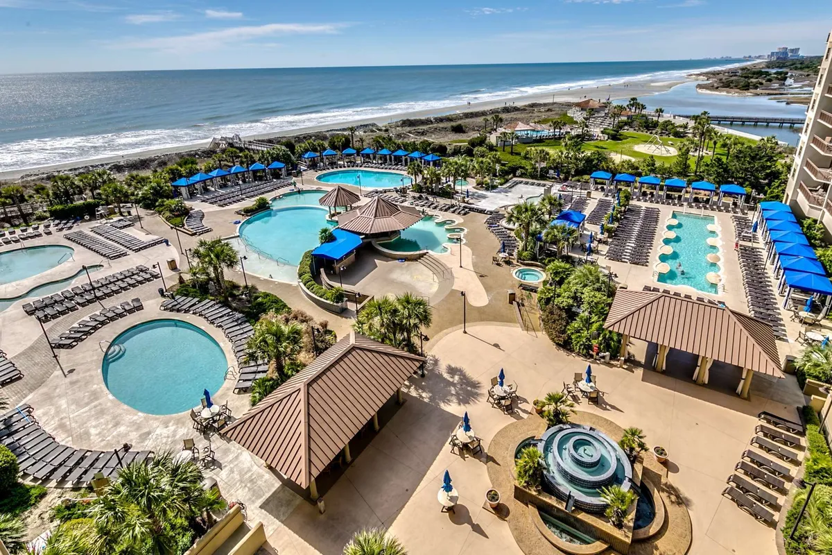 A vibrant exterior scene showcasing a resort-style swimming pool surrounded by tall palm trees, abundant lounge chairs, and shade structures. The beach is visible in the background, reinforcing the proximity to the ocean and the outdoor leisure possibilities. The mood is lively and inviting, suggesting a place where guests can swim, sunbathe, and gather with friends or family.