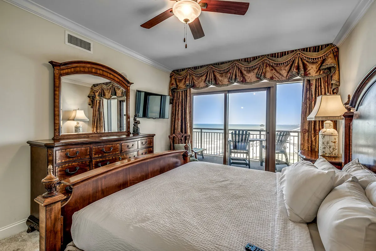 A spacious master bedroom featuring a king-size bed with plush bedding, a large mirror, luxurious dresser, and sliding glass doors revealing an ocean view. The room is bathed in natural light and exudes comfort and tranquility.