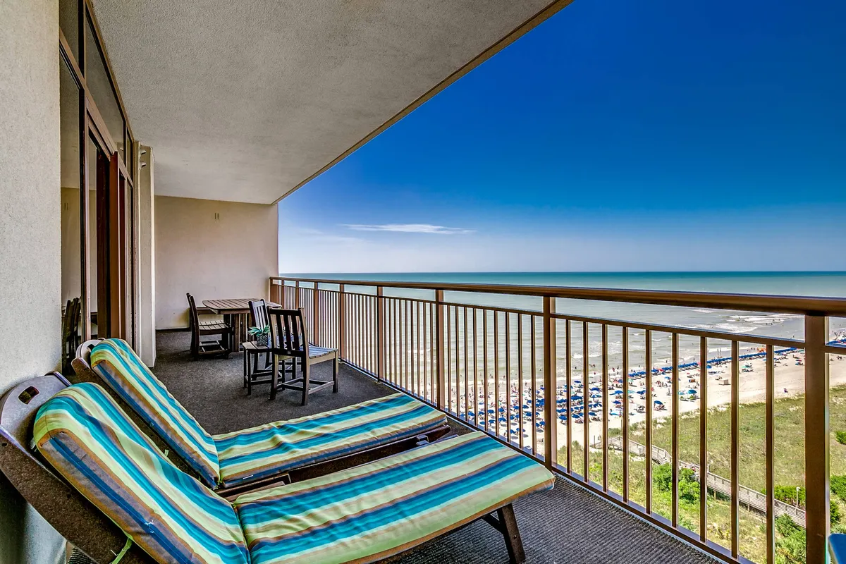 A sunlit private balcony with striped lounge chairs, a balcony railing overlooking a stunning ocean view, and the beach visible below. The setup invites relaxation, from sunbathing to enjoying the ocean breeze and watching the sunset or sunrise.
