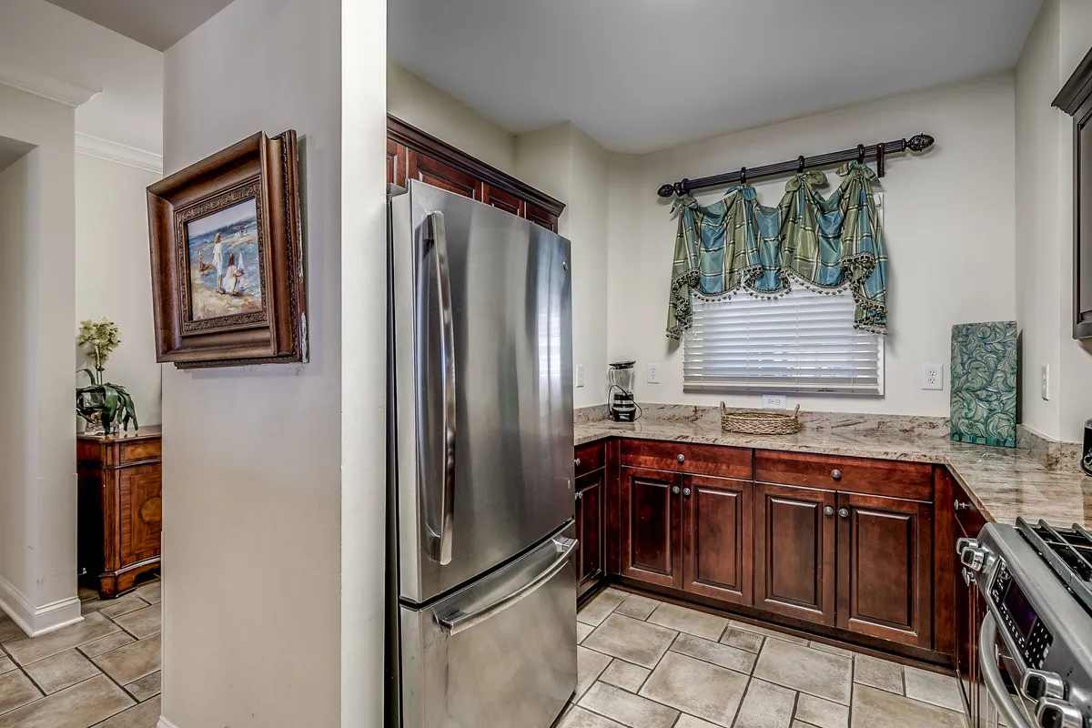 This image captures a spacious kitchen with a stainless steel refrigerator, dark wood cabinets, decorative green curtains, and framed artwork. The setting suggests a practical, welcoming atmosphere ideal for cooking and socializing, with ample space for meal preparation or gathering with family.