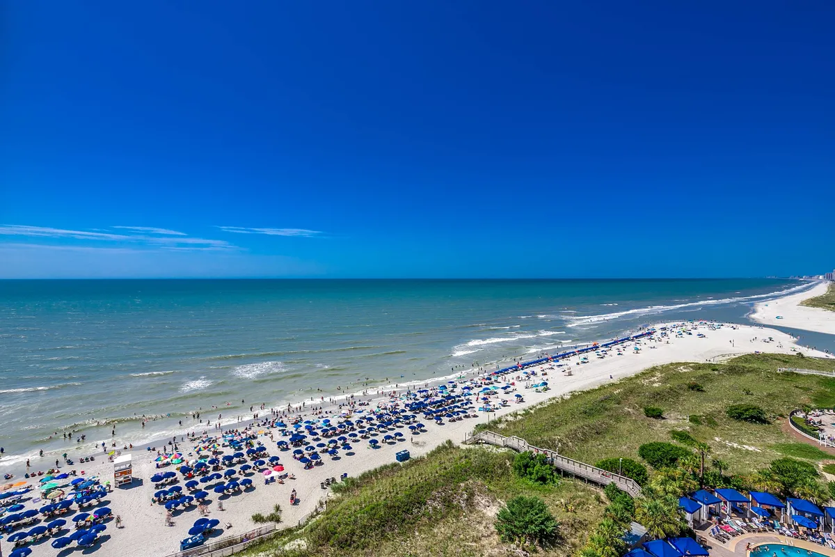 A lively, sunlit stretch of North Myrtle Beach, with families and children playing in the sand, colorful umbrellas dotting the shoreline, and gentle waves rolling in. The scene captures groups building sandcastles, beachgoers tossing balls, and the joyful energy of a family-oriented beach day.