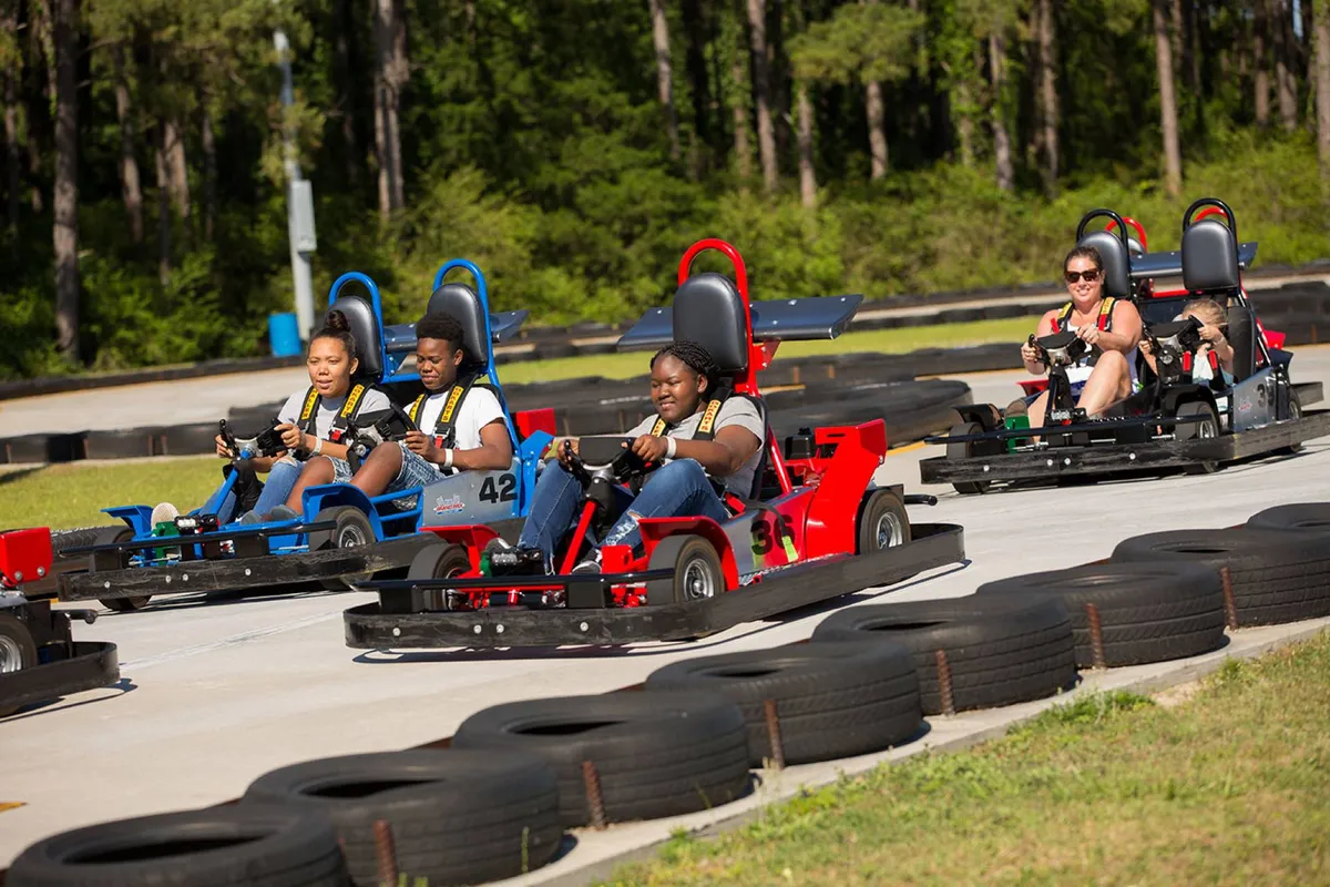 An action-packed shot of families and kids racing go-karts around a winding track, with tire barriers and cheering onlookers in the background. The mood is energetic and playful, capturing the spirit of friendly competition and shared excitement.