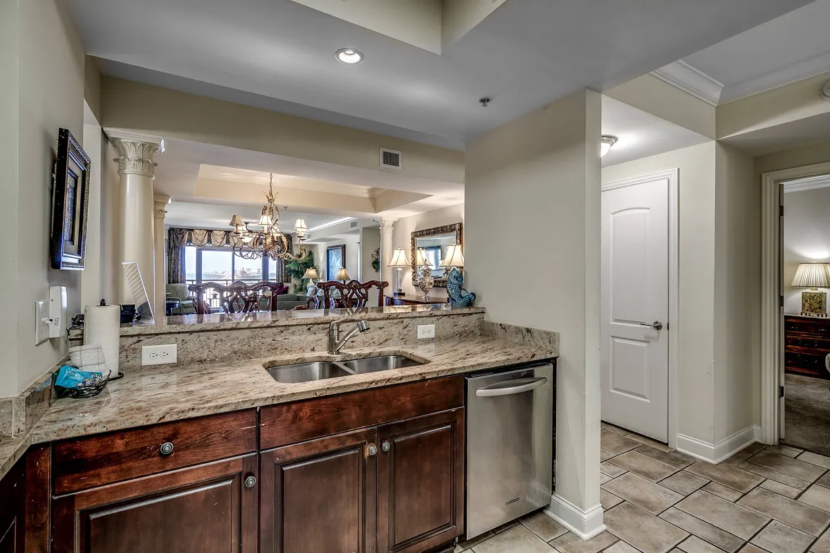 The image presents a sophisticated kitchen view with granite countertops, a double sink, decorative mirror, and a chandelier, with the dining area visible in the background. The elegant setting highlights the seamless flow between kitchen and dining spaces, evoking warmth and togetherness.