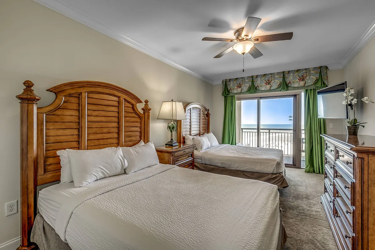 A secondary bedroom with two wooden beds, a dresser, lamp, and access to a balcony. The setting is comfortable and inviting, ideal for friends, families, or couples, with natural light streaming in and tranquil decor.