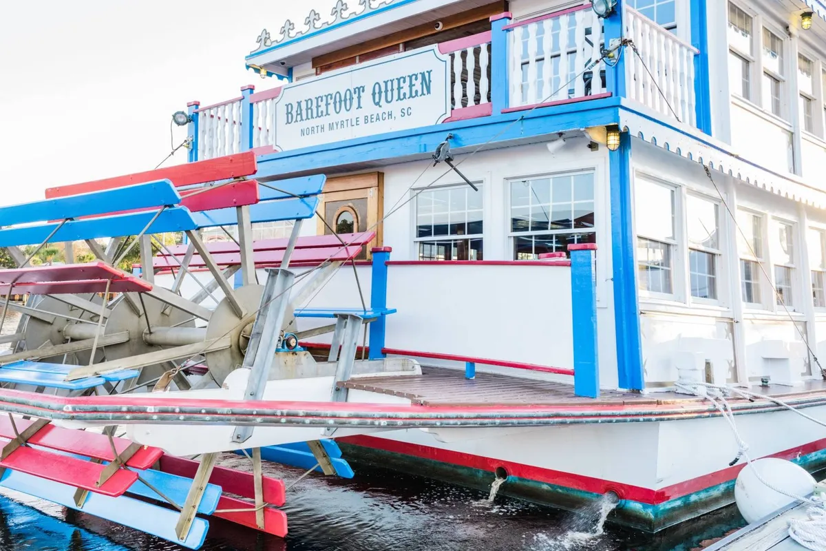 An exterior view of the 'BAREFOOT QUEEN' paddle wheel boat, with its colorful façade and vibrant signage. The scene conveys a cheerful and laid-back mood, evoking the leisure and scenic enjoyment available through water-based activities like cruises and waterfront dining.
