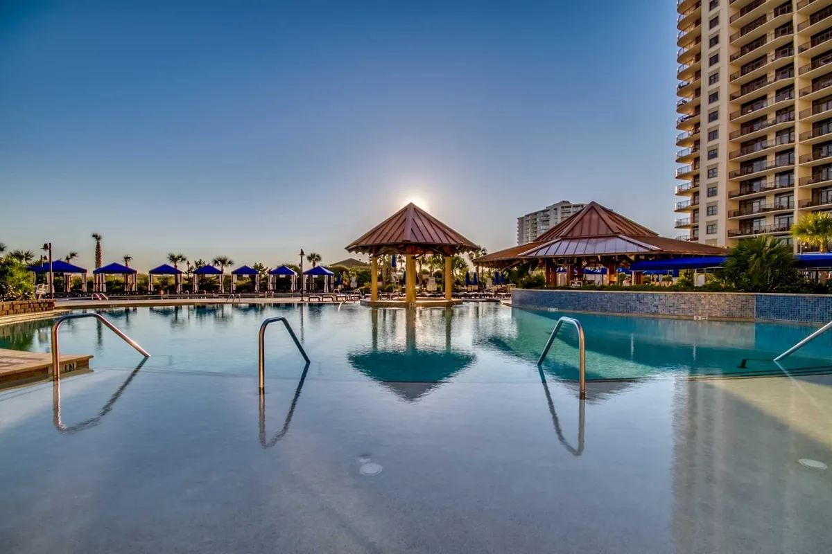 This exterior image features a luxurious swimming pool surrounded by thatched-roof cabanas, with the sun setting in the background. The tranquil atmosphere is enhanced by subtle lighting and palm trees, evoking the high-end, resort-style amenities mentioned in the section.