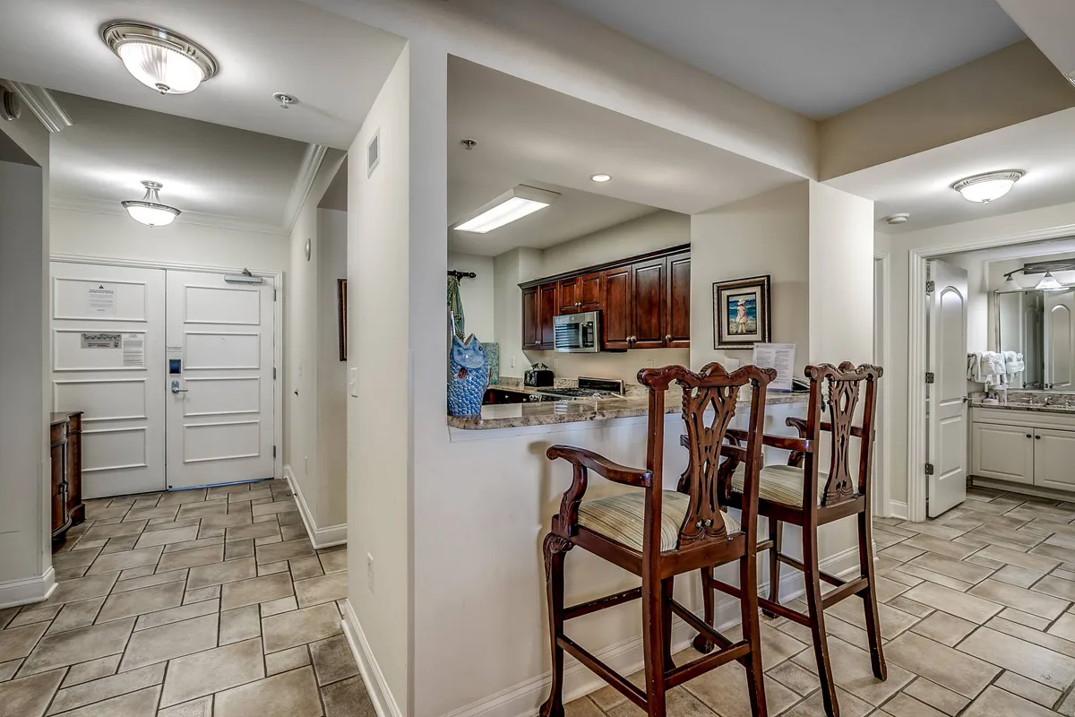 This image shows the open kitchen area with bar stools pulled up to a spacious counter, wooden cabinetry, and the entryway visible in the background. The space is bathed in natural light and designed for both relaxation and socializing, emphasizing a practical yet elegant living experience.