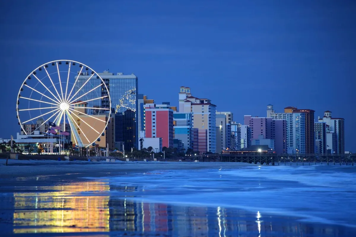 A vibrant exterior scene featuring Myrtle Beach's famous Ferris wheel, colorful buildings, and the shimmering waterfront. The boardwalk atmosphere is lively, hinting at entertainment, nightlife, and the joyful energy of the area after dark.